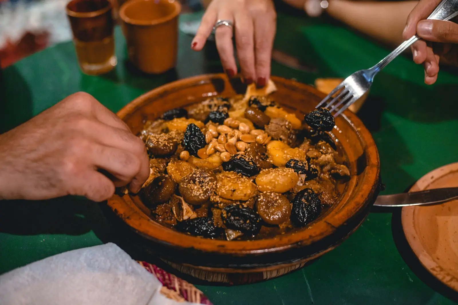 Hands digging into a lamb tagine with prunes and almonds, served in a clay dish